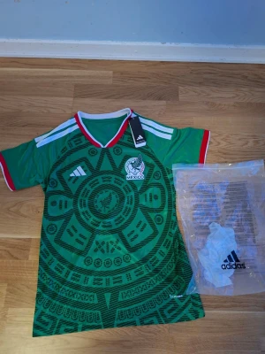 Mexiko matchtröja Adidas grön - Säljer en grön Mexiko matchtröja från Adidas med aztekiskt mönster och vita detaljer. Tröjan har röd och vit V-ringad krage, samt Mexikos landslagsemblem på bröstet. Tillverkad i lätt och ventilerande material, perfekt för fotboll eller träning.