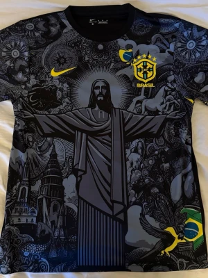 Nike Brasil grafisk fotbollströja - Unik svart fotbollströja från Nike med Brasil-logga och gult Swoosh. Framsidan har ett stort grafiskt tryck med Kristusstatyn, brasilianska symboler och detaljer i gult. Tillverkad i lätt och ventilerande material, perfekt för träning eller match.