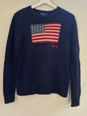 Mörkblå stickad tröja Polo Ralph Lauren - Mörkblå stickad tröja från Polo Ralph Lauren med amerikansk flagga i rött, vitt och blått på bröstet samt broderade initialer 'RL'. Tröjan har rund halsringning, ribbade muddar och är tillverkad i mjukt garn. Perfekt för en klassisk och avslappnad look.
