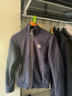 Moncler zip up - Mörkblå ziptröja från Moncler med hög krage och dragkedja framtill. Tröjan har två fickor med randiga detaljer och Moncler-logga på bröstet. Tillverkad i mjukt material som känns bekvämt och stilrent. Storlek M, priset är ej hugget i sten!