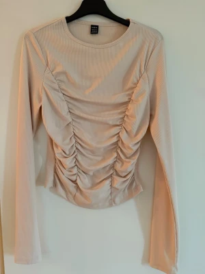 Beige ribbad topp från SHEIN - Trendig beige långärmad topp från SHEIN med ribbad struktur och snygga rynkade detaljer framtill. Figurnära passform som framhäver midjan och ger en modern look. Perfekt att styla med jeans eller kjol för en cool outfit.