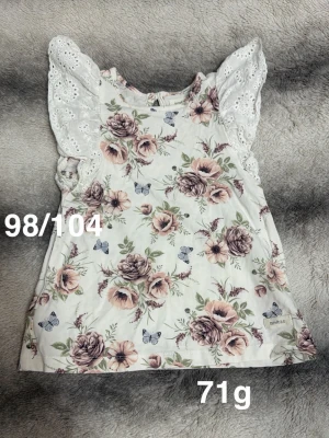 Vit blommig topp med spetsvolang - Romantisk topp i vitt med blommigt mönster i rosa och lila toner samt små fjärilar. Ärmlös modell med breda spetsvolanger på axlarna som ger en söt och feminin look. Perfekt för sommarens alla tillfällen.🌸Fint använt skick. Storlek 98/104