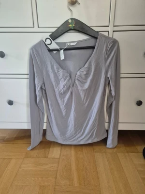 Ljusgrå långärmad topp med v-ringning - Säljer en helt ny ljusgrå långärmad topp med snygg v-ringning och rynkade detaljer framtill. Toppen har en smickrande passform och är perfekt att styla till jeans eller kjol.