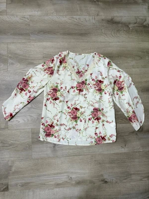 Vit blommig blus med v-ringning - Söt vit blus med rosa och röda blommor samt v-ringning. Blusen har trekvartsärm och ett romantiskt blommönster över hela plagget. Perfekt för dig som gillar en feminin och fräsch stil. Materialet känns lätt och luftigt.