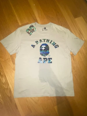 Vit BAPE t-shirt med blått camotryck - Säljer en clean vit t-shirt från A Bathing Ape med ikoniskt blått och grönt camotryck på bröstet och stor BAPE-logga. Klassisk passform och rund hals. Perfekt för dig som vill sticka ut med streetwear-vibe.