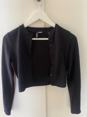 Svart kort kofta  -  Modellen är kort och har långa ärmar, perfekt att slänga över en topp. Materialet ser mjukt och bekvämt ut, och koftan har en enkel, klassisk design.