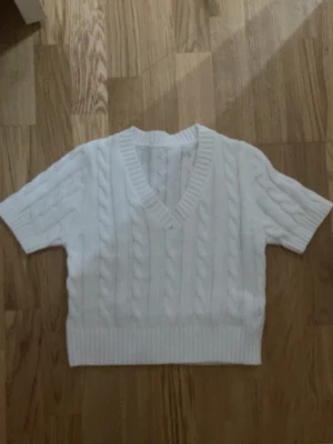 Vit kabelstickad kortärmad tröja - Säljer en vit kabelstickad tröja med korta ärmar och djup V-ringning. Tröjan har bred ribbad kant nedtill och vid ärmsluten, vilket ger en snygg och modern look. Perfekt att styla med jeans eller kjol för en trendig outfit.