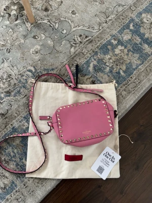 Rosa axelväska Valentino med nitar - Säljer en rosa axelväska från Valentino med guldfärgade nitar runt kanterna. Väskan har justerbar axelrem och är tillverkad i slätt läder. Perfekt för dig som vill ha en statementväska med edgy detaljer och exklusiv känsla.