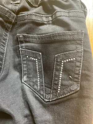 Svarta jeans med nitar på bakfickan - Snygga svarta jeans med raka ben och dekorativa silvriga nitar på bakfickan. Jeansen har klassisk femficksdesign och är tillverkade i ett stretchigt denimtyg som sitter bekvämt. Perfekta för dig som gillar en stilren look med lite extra detaljer.