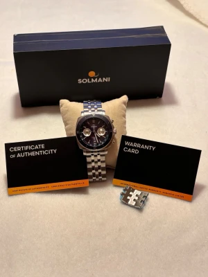 Solmani silverfärgad kronografklocka - Stilren kronografklocka från Solmani med silverfärgad länkarmband i metall och svart urtavla. Klockan har två subdials, datumvisning och markerad bezel. Levereras med originalask, certifikat och extra länk.