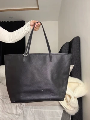 Stor svart shoppingväska i skinnimitation - Snygg och rymlig svart shoppingväska med långsmala handtag. Väskan har en stilren, enkel design och är gjord i skinnimitation med lätt glansig yta. Perfekt för att bära allt du behöver till skolan, jobbet eller stan.