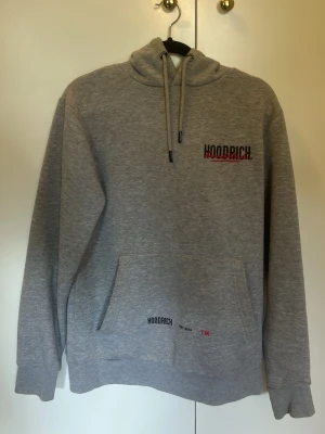 Grå hoodie från Hoodrich - Snygg grå hoodie från Hoodrich med broderad logga i svart och rött på bröstet. Klassisk känguruficka framtill och justerbar huva med dragsko. Tillverkad i mjukt bomullsmaterial som är perfekt för chill dagar. Hoodrich-detaljer även på fickan.