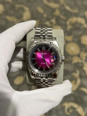 Seiko mod automatisk klocka silver/rosa - Snygg automatisk klocka från Seiko med metallarmband i silver och en unik rosa urtavla. Klockan har datumvisning och klassisk räfflad bezel. Perfekt accessoar för dig som vill sticka ut med stilren design och cool färgkombination.