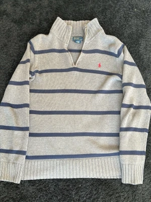 Gråblå randig stickad tröja Polo Ralph Lauren - Strl L i barn storlek Stickad tröja från Polo Ralph Lauren i ljusgrått med mörkblå horisontella ränder och rosa logga på bröstet. Tröjan har halv dragkedja vid halsen, ribbade muddar och är långärmad. Perfekt för en avslappnad men snygg look.