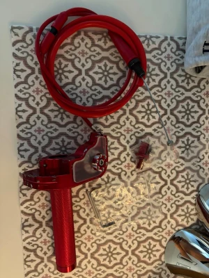 Röd snabbgas till moped - Säljer ett rött gasreglage med matchande gasvajer och tillbehör. Handtaget är i metall med greppvänlig yta och transparent plastdetalj. Perfekt för dig som vill uppgradera din moped eller scooter med en snygg och sportig look.