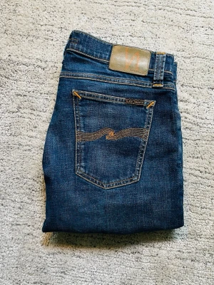 Nudie jeans - Säljer ett par klassiska blå jeans från Nudie Jeans med snygga slitningar framtill och ikonisk bakficka med vågig söm. Jeansen har normal midja, smal passform och är gjorda i mjuk denim med kontrastsömmar. Perfekta för en avslappnad och trendig look.
