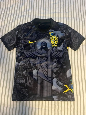 Nike Brasil Jesus svart matchtröja - Unik svart Brasilien-matchtröja från Nike med detaljerat grafiskt tryck av Kristusstatyn, kyrkor och brasilianska symboler. Gula loggor och emblem på bröstet, kort ärm och rund hals. Perfekt för dig som vill sticka ut på planen eller läktaren.