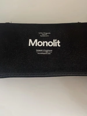 Monolit svarta kolfiberbenskydd - Säljer ett par svarta benskydd från Monolit i kolfiber med ergonomisk form och vadderad insida. De levereras i en svart förvaringsväska med dragkedja. Perfekta för dig som vill ha lätta och slitstarka skydd till sport.