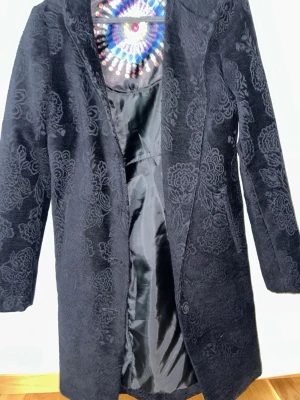 Svart mönstrad kappa från Desigual - Snygg svart kappa från Desigual med diskret blommönster i sammet och färgglatt foder. Rak modell med långa ärmar och öppen front. Perfekt för dig som vill ha en unik och stilren look med en twist. Materialet känns mjukt och lyxigt.
