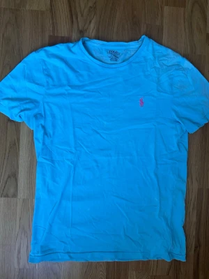 Blå t-shirt från Polo Ralph Lauren - Snygg blå t-shirt från Polo Ralph Lauren med klassisk rund hals och rosa broderad logga på bröstet. Modellen är custom slim fit och tillverkad i mjuk bomull, perfekt för en avslappnad och stilren look. Det finns en lite defekt som ni ser på 3 e bilden men förutom det är det chill