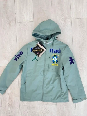 Brasil vindjacka med huva GORE-TEX - Ljusgrön vindjacka från Jordan med Brasil CBF-logga, GORE-TEX-material och huva. Jackan har tryckta sponsorloggor i blått och grönt samt dragkedja framtill. Perfekt för dig som gillar sportig stil och vill hålla dig torr. Står M men passar S-XS