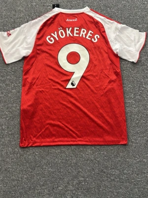 Arsenal matchtröja Gyökeres #9 - Snygg Arsenal matchtröja från Adidas i rött och vitt med Gyökeres och nummer 9 på ryggen. Tröjan har klubbmärke, Adidas-logga och sponsortryck framtill samt klassiska ränder på axlarna. Tillverkad i lätt och ventilerande material, perfekt för fotboll eller träning.