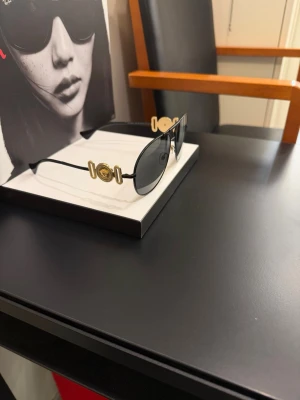 Svarta solglasögon från Versace - Snygga svarta solglasögon från Versace med klassisk aviatorform. Tunn metallbåge i svart med guldiga detaljer och ikonisk Medusa-logga på sidorna. Perfekta för dig som vill ha en lyxig och trendig accessoar till sommaren.
