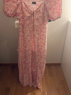 Blommig långklänning Gina Tricot - Långklänning från Gina Tricot i ett blommigt mönster med rosa, vita och gröna toner. Klänningen har puffärmar, knytning framtill och en feminin siluett. Perfekt för sommarens alla tillfällen.