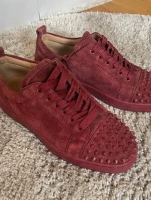 Bordeauxröda sneakers från Louboutin - Säljer ett par bordeauxröda sneakers från Christian Louboutin i mocka med matchande snören och coola nitar på tån. Skorna har en låg silhuett och en chunky sula som ger en riktigt lyxig vibe. Perfekta för dig som vill sticka ut med exklusiv design.