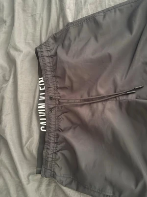 Svarta badbyxor från Calvin Klein - Snygga svarta badbyxor från Calvin Klein med resårmidja och justerbar dragsko. Tydlig logga på insidan av midjan i vitt. Tillverkade i snabbtorkande material, perfekta för stranden eller poolen.