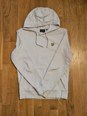 Vit hoodie från Lyle & Scott - Snygg vit hoodie från Lyle & Scott med klassisk gul logga på bröstet. Hoodien har huva med dragsko och en stor magficka framtill. Tillverkad i mjukt material som känns skönt mot huden. Perfekt för en avslappnad och stilren look.