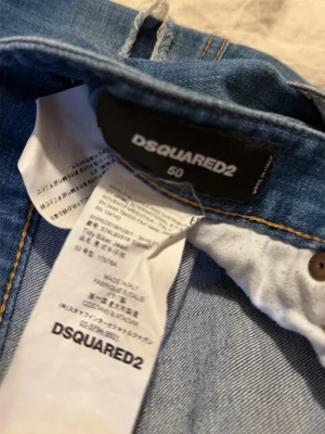 Blå jeans från DSQUARED2 med patch - Säljer ett par blå jeans från DSQUARED2 i storlek 50. Jeansen har en unik färgglad patch med motiv bak och coola slitningar samt kontrastsömmar. Klassisk femficksmodell i bomull med detaljer som sticker ut. Perfekta för dig som vill ha något extra.