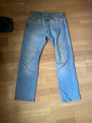 Levis 501 Jeans - Klassiska ljusblå jeans från Levi's med rak passform och fem fickor. Jeansen har knappgylf och tydliga slitningar för en vintagekänsla. Tillverkade i denim av bomull. Perfekta för en avslappnad och trendig look. W32 L32