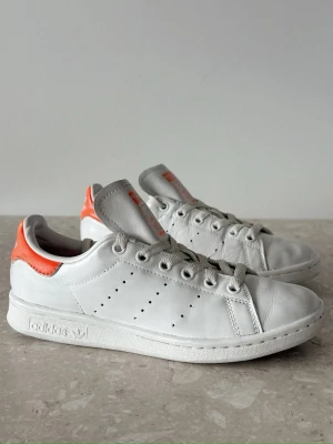 Adidas Stan Smith | EU 37.5  - Klassiska Adidas Stan Smith sneakers i vitt läder med orange detaljer på hälen och tungan. Skorna har perforerade ränder på sidorna, platt sula och snörning framtill. Perfekt för dig som gillar en clean och tidlös look.
