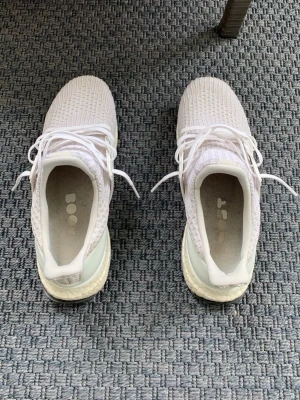 Vita Adidas Ultraboost  - Säljer ett par vita Adidas Ultraboost sneakers med stickad ovandel och klassiska vita snören. Skorna har en bekväm Boost-sula och diskret Adidas-logga på hälen. Perfekta för dig som gillar stilrena och sportiga sneakers.