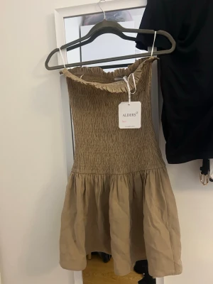 Beige smockad klänning från ALDERS - Supersöt beige klänning från ALDERS med smockad överdel och volangkant upptill. Klänningen är axelbandslös och har en luftig kjol i lätt material, perfekt för varma dagar. Enkel och trendig design som passar till många tillfällen.