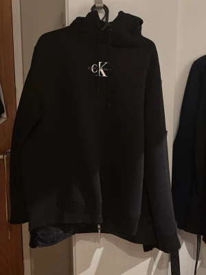 Svart hoodie från Calvin Klein - Snygg svart hoodie från Calvin Klein med klassisk CK-logga på bröstet. Hoodien har huva med dragsko och ribbade muddar vid ärmslut och nederkant. Perfekt för en chill och stilren look.
