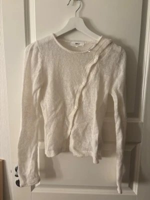 Vit kofta med knappar från Gina Tricot - Säljer en vit, mjuk kofta från Gina Tricot med lång ärm och rund hals. Koftan har en unik knäppning med små vita knappar som går snett över framsidan och ribbade detaljer vid kanterna. Perfekt för lager-på-lager och enkel att matcha.