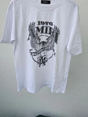 Vit Amiri t-shirt med örntryck - Snygg vit t-shirt från Amiri med stort svart tryck framtill som visar en örn, texten '1976' och 'Hollywood California'. Klassisk rund hals och korta ärmar. Perfekt för dig som gillar streetwear och vill sticka ut med ett coolt motiv.