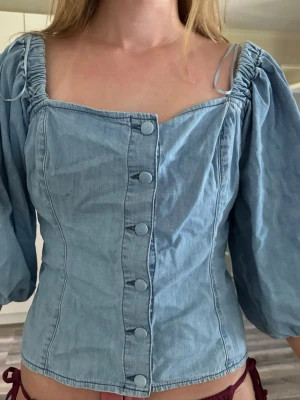 Ljusblå denim blus med puffärm - Trendig ljusblå blus i denim-look med puffiga trekvartsärmar och knappar framtill. Fyrkantig ringning och figurnära passform ger en snygg siluett. Perfekt till jeans eller kjol för en cool vardagsstil.