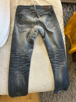 Blå slitna jeans från Nudie Jeans - Säljer ett par blå jeans från Lee med tydliga slitningar och tvättade detaljer. Jeansen har raka ben, klassiska bakfickor och en patch i läder bak i midjan. Materialet är denim i bomull och passformen är smal med snygga whiskers-effekter.