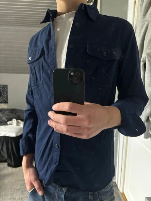 Mörkblå Overshirt - Snygg mörkblå Overshirt med klassisk krage och knappar framtill. Två bröstfickor med lock och knappdetaljer ger en stilren look. Skjortan har lång ärm och är tillverkad i ett mjukt bomullsmaterial. Perfekt för en avslappnad men ändå uppklädd stil.