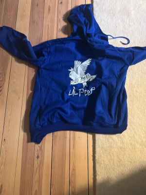 Blå hoodie med Lil Peep-tryck - Säljer en blå hoodie med stor huva och tryck av en vit duva samt texten 'Cry Baby' och 'Lil Peep' på ryggen. Hoodien har ribbade muddar och en avslappnad passform, perfekt för dig som gillar streetwear och vill sticka ut.