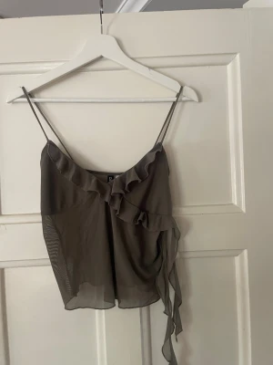 Militärgrön volang topp från Hm - Snygg grön linnetopp från hm med tunna axelband och volangdetaljer framtill. Toppen har omlottkänsla och är gjord i ett tunt, transparent material som känns luftigt och lätt.