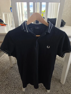 Svart pikétröja från Fred Perry - Klassisk svart pikétröja från Fred Perry med vit broderad logga på bröstet och vita ränder på krage och ärmslut. Tröjan har korta ärmar och en stilren, sportig look som passar perfekt till jeans eller shorts.