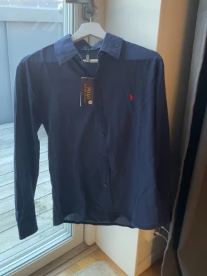Mörkblå skjorta från Polo Ralph Lauren - Klassisk mörkblå skjorta från Polo Ralph Lauren med lång ärm och knappar framtill. Skjortan har den ikoniska röda logotypen broderad på bröstet och är tillverkad i mjuk bomull. Perfekt för en clean och stilren look. Den var för liten så säljer den 