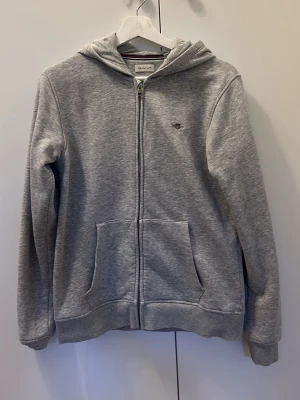 Grå zip hoodie från GANT - Snygg grå hoodie från GANT med dragkedja framtill och två stora fickor. Klassisk huva och liten broderad logga på bröstet. Tillverkad i mjuk bomullsblandning, perfekt för chill dagar eller när du vill ha en clean look.