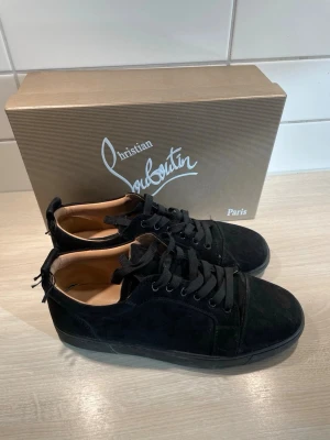 Svarta Christian Louboutin sneakers - Snygga svarta sneakers från Christian Louboutin i mocka med klassiska röda sulor och svarta skosnören. Låg modell med stilren design och exklusiv känsla. Perfekta för dig som vill ha något extra till din outfit. Box ingår!