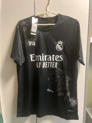 Svart Real Madrid matchtröja Y-3 - Snygg svart Real Madrid matchtröja i samarbete med Y-3 och Adidas. Tröjan har vita tryck med klubbmärke, sponsor och Y-3-logga framtill samt diskreta rosdetaljer. Tillverkad i lätt och funktionellt material, perfekt för fotboll eller träning.