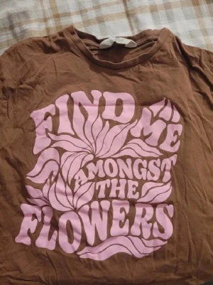 Brun t-shirt med rosa tryck - Cool brun t-shirt med stort retroinspirerat rosa tryck på framsidan där det står 'Find me amongst the flowers' i vågig text och blommönster. Perfekt för dig som gillar vintage vibes och statement-plagg.